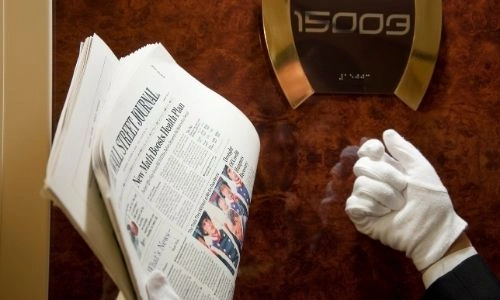 main d’un majordome qui frappe à la porte d’une suite, tenant dans ses mains des journaux