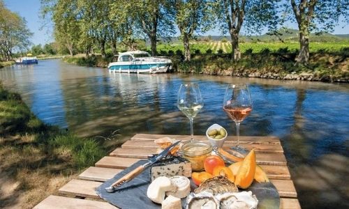 présentation d’un plateau de dégustation sur un ponton d’un bord de rivière au premier plan avec un bateau sans permis au second plan