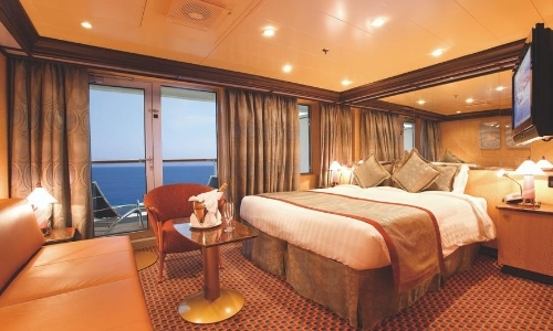 Vue de l'intérieur d'une chambre sur un navire de croisière avec grand lit, fauteuil, canapé, TV, et fenêtres avec vue sur la mer