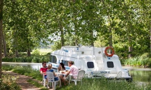famille qui profite d’un repas sous les arbres sur le bord d’un fleuve à côté de leur péniche 