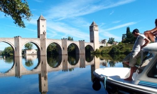 vue d’un magnifique pont à arcs et de l’avant d’une péniche sur laquelle est adossé un homme et une femme sur le toit 