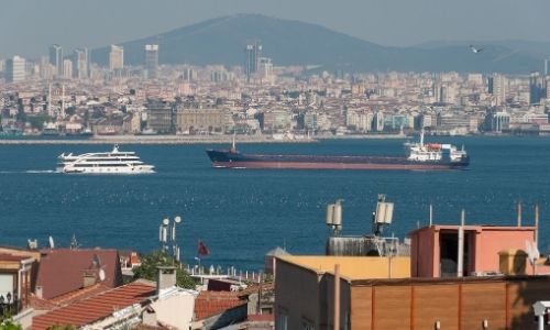 premier plan avec des habitations, second plan la mer avec des cargo dessus et un arrière-plan de la ville et de montagnes au loin