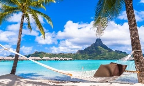 hamac au premier plan accroché à des palmiers sur le sable avec la mer turquoise en paysage et des montagnes au loin