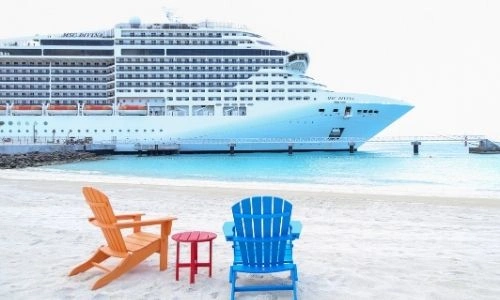 une chaise en bois peinte en orange et une autre en bleue, accompagnées d’une petite table rouge sur le sable avec un bateau MSC sur l’eau en arrière-plan.