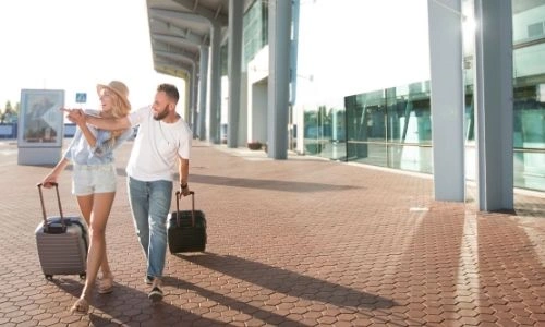 un couple en dehors de l&rsquo;a&eacute;roport, roulent des valises et indiquent avec leurs mains entrelac&eacute;es une direction 