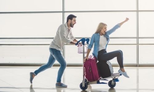 un jeune homme pousse une jeune femme assise sur un porte valise dans un aéroport avec enthousiasme