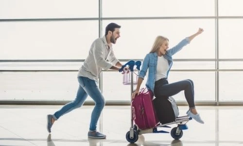 un jeune homme pousse une jeune femme assise sur un porte valise dans un a&eacute;roport avec enthousiasme