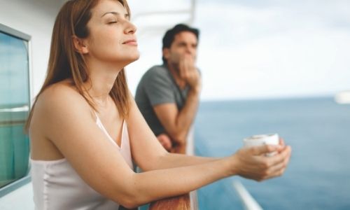 jeune femme qui respire le grand air sur le balcon d’une croisière et jeune homme en arrière-plan qui observe la mer, tous deux, adossé à la barre