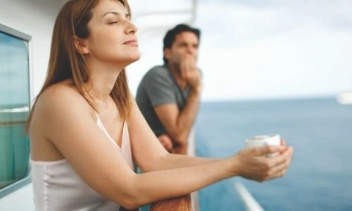 jeune femme qui respire le grand air sur le balcon d&rsquo;une croisi&egrave;re et jeune homme en arri&egrave;re-plan qui observe la mer, tous deux, adoss&eacute; &agrave; la barre
