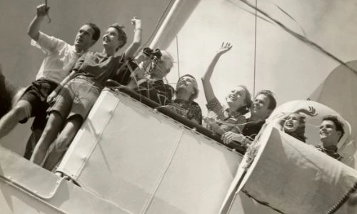 photo en noir et blanc d’un groupe de personnes qui sont à bord d’un bateau, et disent au-revoir d’un signe de la main, avec enthousiasme