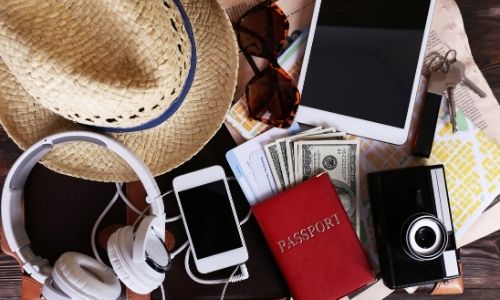 accessoires pour partir en vacances étalés sur un lit : casque, téléphone, passeport, appareil photo, billets, tablette, lunettes de soleil