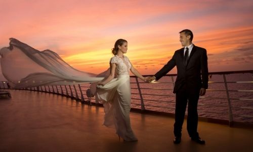 couple qui se regardent et se tiennent la main, habillés de manière classe avec la robe de la dame qui prend le vent, sur le pont d’un bateau 