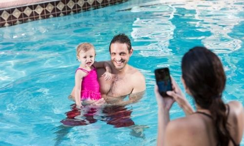 jeune femme de dos qui tient dans ses mains un téléphone pour prendre son homme et sa petite fille en photo dans la piscine, eux sont en face d’elle et sourient