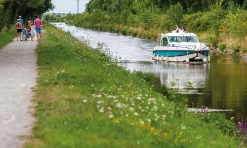 Famille qui se promène le long d’un canal, sur lequel on retrouve une pénichette