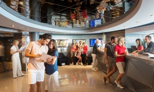 hall d’entrée d’un bateau de croisière avec beaucoup de personnes qui se renseignent aux quatres coins du hall, guichets, canapés, etc