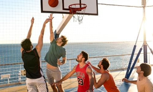 cinq jeunes garçons qui jouent au basketball sur un terrain multisport d’une croisière, l’un est en train de shooter pendant qu’un autre défend sous le regard des trois autres