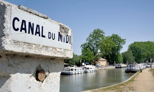 Le canal du Midi et ses péniches blanches. Un panneau intitulé “Canal du Midi” se trouve en premier plan