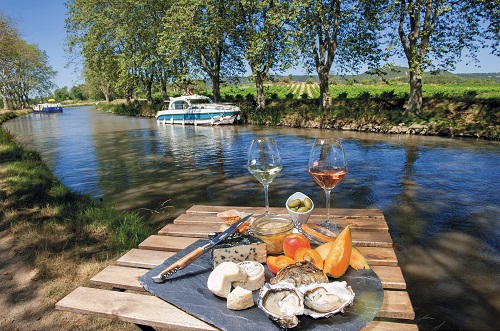Table de pique nique terroir (fromages, huîtres, fruits et verres de vin) devant le canal du Midi et ses platanes. Un bateau de location est amarré sur une berge. 