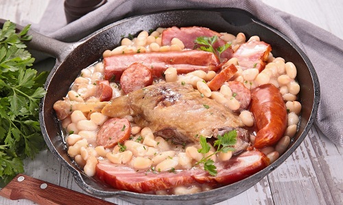 Du cassoulet composé de haricots blancs, du confit de canard, du jarret de porc de la saucisse et du persil présenté dans une poêle à sur une table blanche