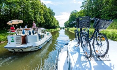 deux péniches sur un canal, l’un peuplé de personnes qui profitent du soleil, l’autre qui porte sur son toit des bicyclettes