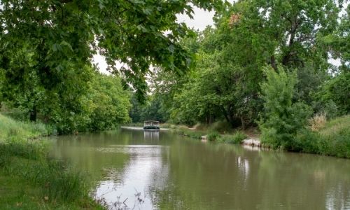 Une péniche navigue sur le Canal du Midi, la nature alentour berce la photo dans un climat de tranquillité