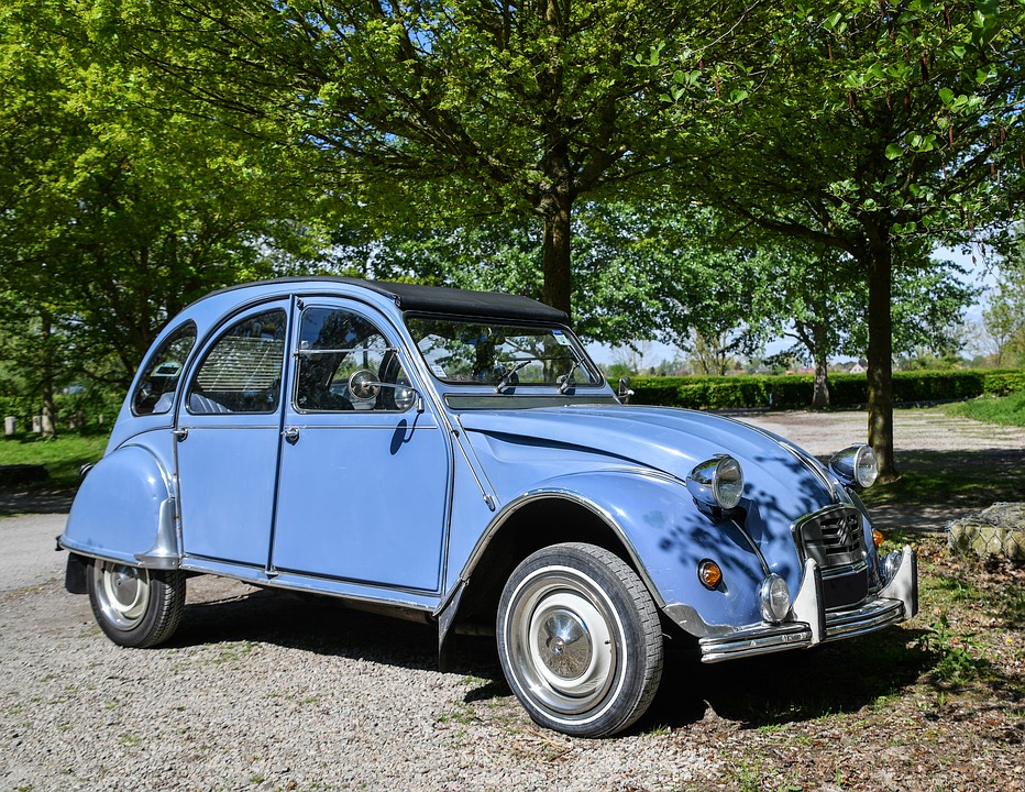 Une 2 chevaux bleue est garée sur un chemin de gravier proche d’arbres.